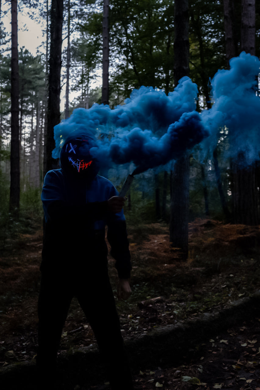 Masked figure holding a blue smoke flare in a forest